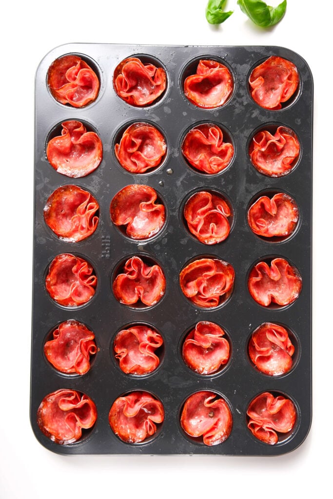 Molde para mini muffins lleno de tazas de salami horneadas, crujientes y listas para rellenar y hacer un aperitivo sabroso.