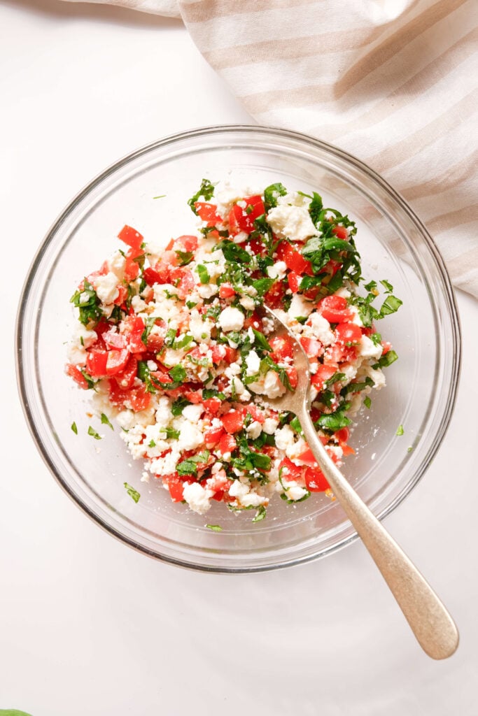 Un tazón de vidrio lleno de una mezcla de tomates picados, feta desmenuzado y albahaca fresca, con una cuchara lista para servir.