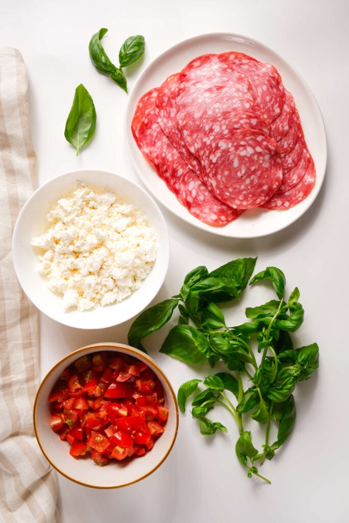 Ingredientes para los bocados de salami crujientes dispuestos sobre una superficie blanca, incluyendo lonchas de salami en un plato, feta desmenuzado en un tazón, tomates picados en un tazón y hojas de albahaca fresca.