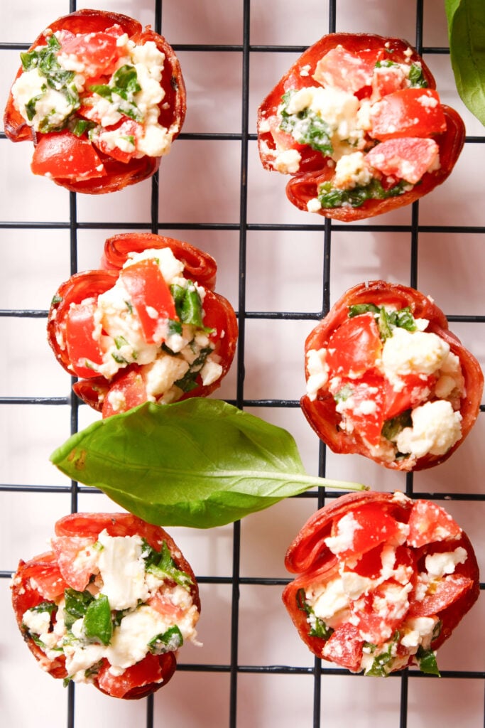 Bocados de salami crujientes rellenos de una mezcla de tomate, feta y albahaca dispuestos sobre una rejilla de enfriamiento con hojas de albahaca fresca.
