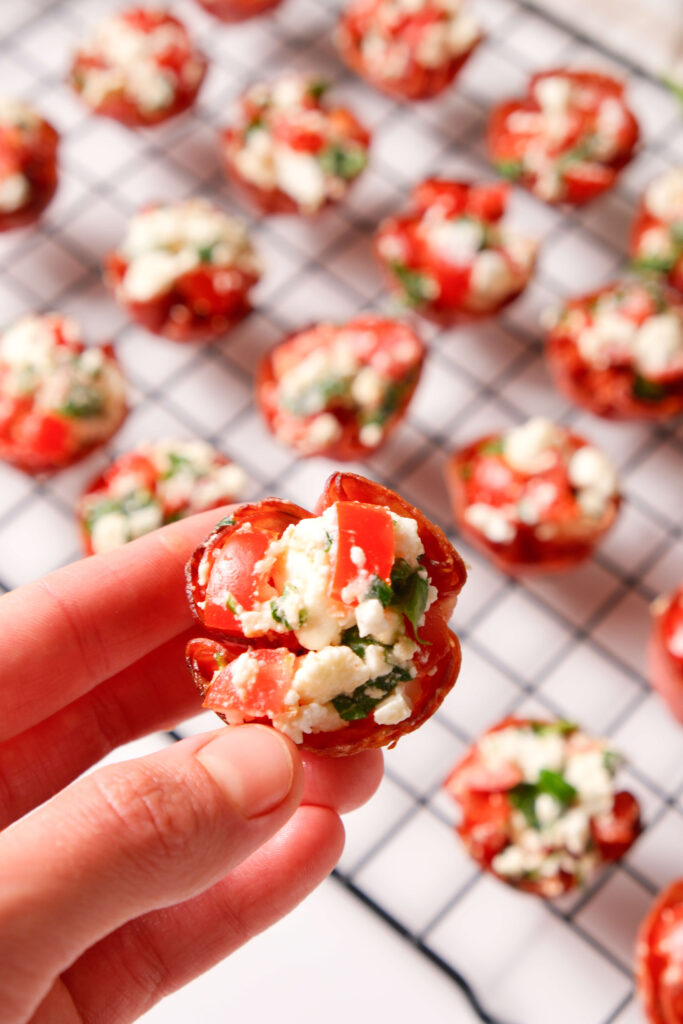 Primer plano de una mano sosteniendo un bocado de salami crujiente relleno de feta, tomate y albahaca, con más bocados de salami en una rejilla de enfriamiento al fondo.