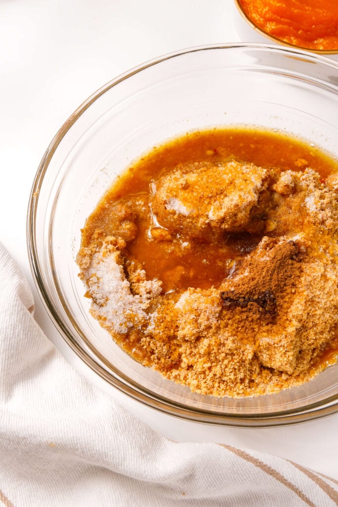 Tazón con migas de galleta graham, azúcar moreno, canela y mantequilla derretida mezclándose para la base de un cheesecake de calabaza.
