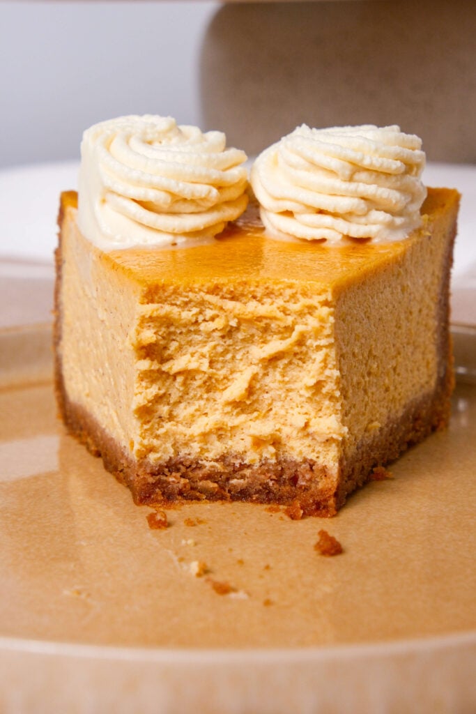 Un primer plano de una porción de cheesecake de calabaza con un bocado tomado, coronada con remolinos de crema batida de canela, mostrando la textura cremosa y la rica base de galleta graham.