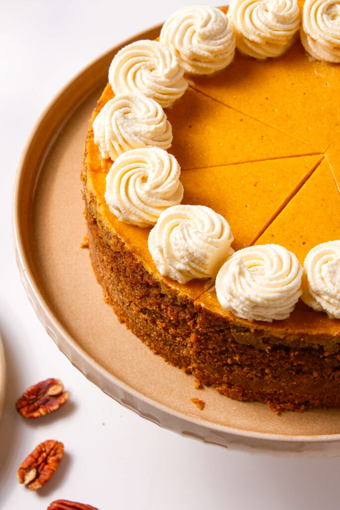 Un primer plano de un cheesecake de calabaza cortado, coronado con remolinos de crema batida de canela, mostrando la base detallada y la textura suave.