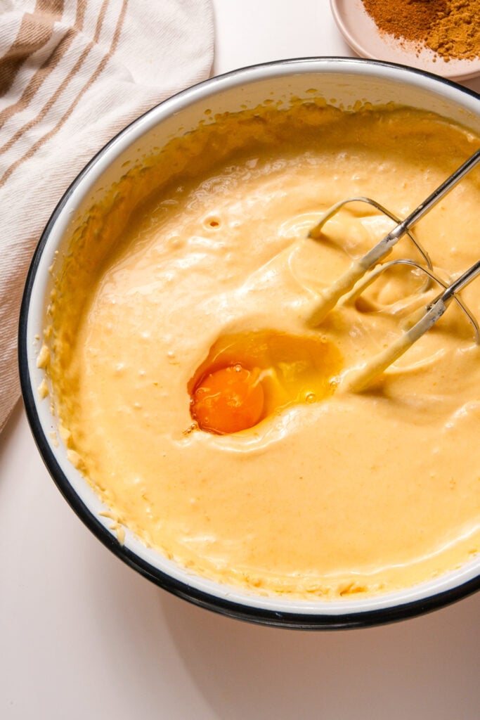 Un huevo agregado a la mezcla de cheesecake de calabaza en un tazón, con una batidora de mano lista para mezclar.