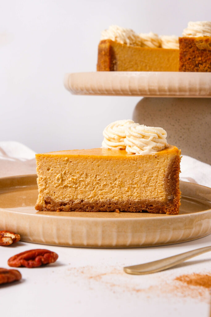 Una vista lateral de una porción de cheesecake de calabaza en un plato, con una base de galleta graham y coronada con remolinos de crema batida de canela, con el cheesecake completo visible en el fondo.