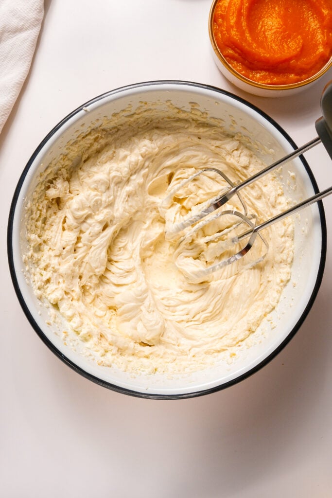 Tazón de queso crema y azúcares batidos con una batidora de mano hasta que estén esponjosos para la base de un relleno de cheesecake de calabaza cremoso.