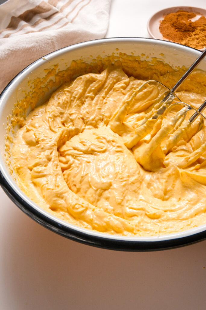 Tazón de mezcla del relleno de cheesecake de calabaza batido con una batidora de mano, incluyendo puré de calabaza, crema agria, extracto de vainilla y jugo de limón.