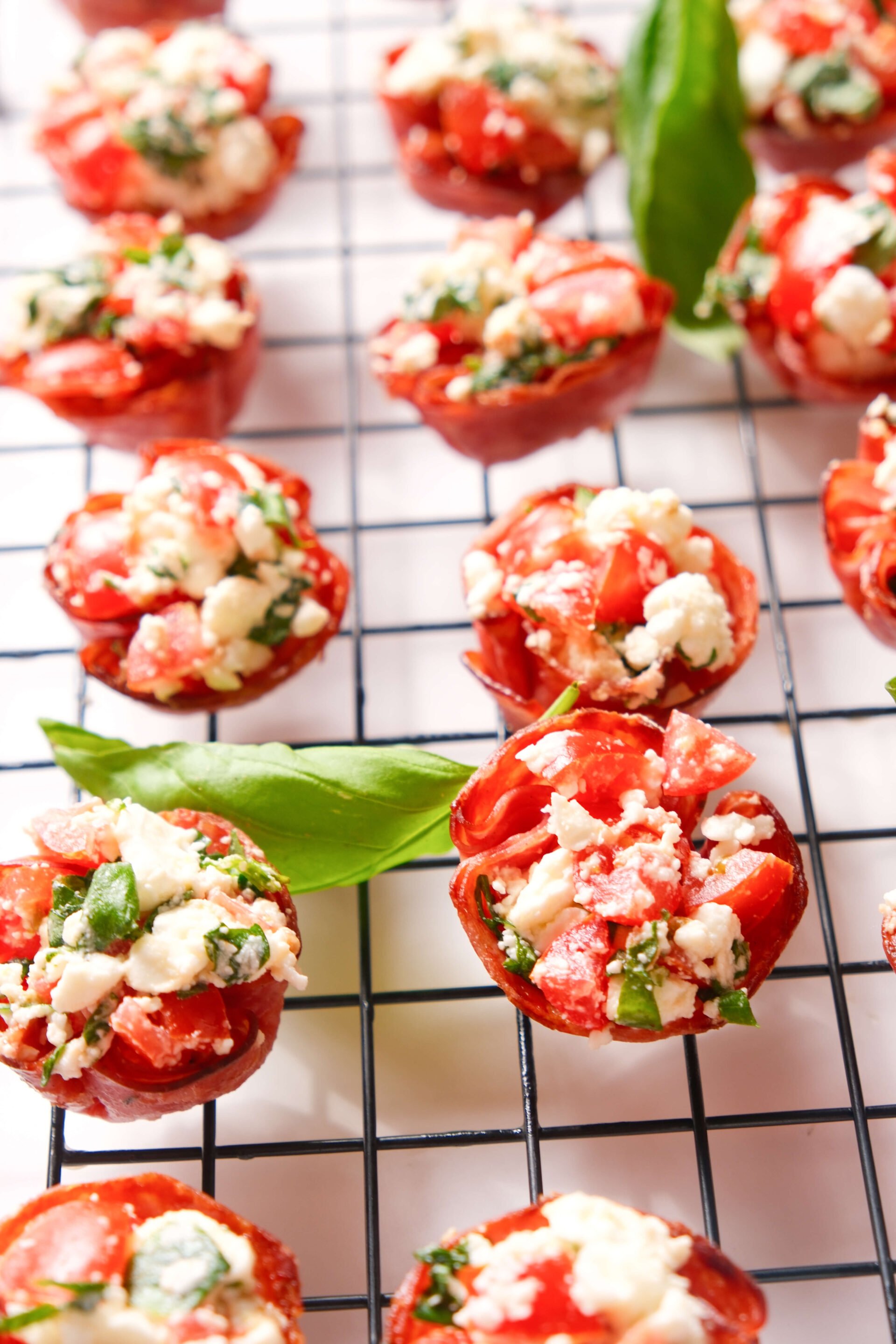 Bocados de salami crujientes rellenos de feta, tomate y albahaca, dispuestos sobre una rejilla para enfriar, decorados con hojas de albahaca fresca.