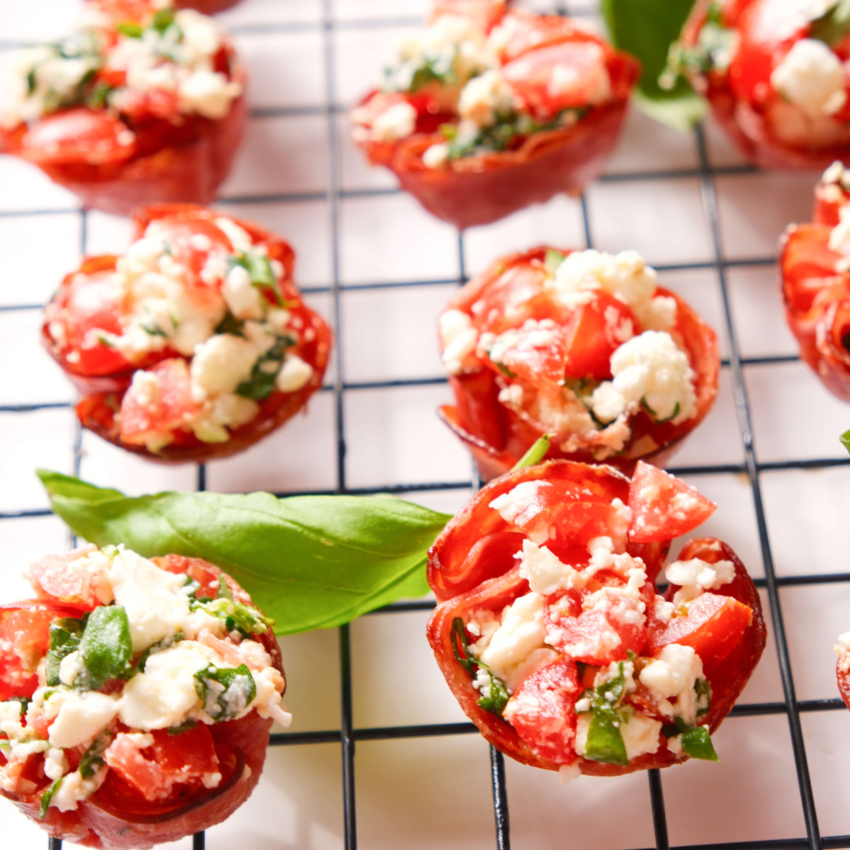 Bocados de salami crujientes rellenos de feta, tomate y albahaca dispuestos sobre una rejilla para enfriar, decorados con hojas de albahaca fresca.