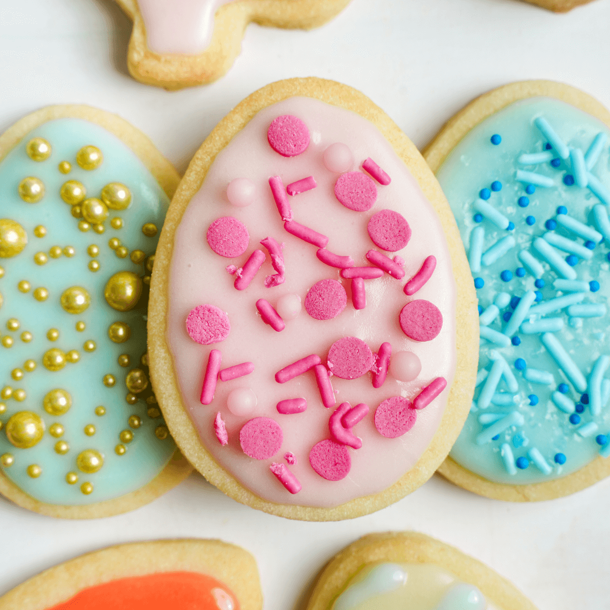 Galleta de azúcar en forma de huevo de Pascua decorada con glaseado rosa claro y chispas rosas, rodeada de galletas de colores pastel en azul, verde y naranja con diversas chispas.