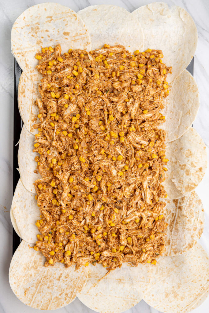 Relleno de quesadillas de pollo al horno en bandeja con pollo desmenuzado y maíz distribuido uniformemente sobre tortillas de harina superpuestas en una bandeja para hornear