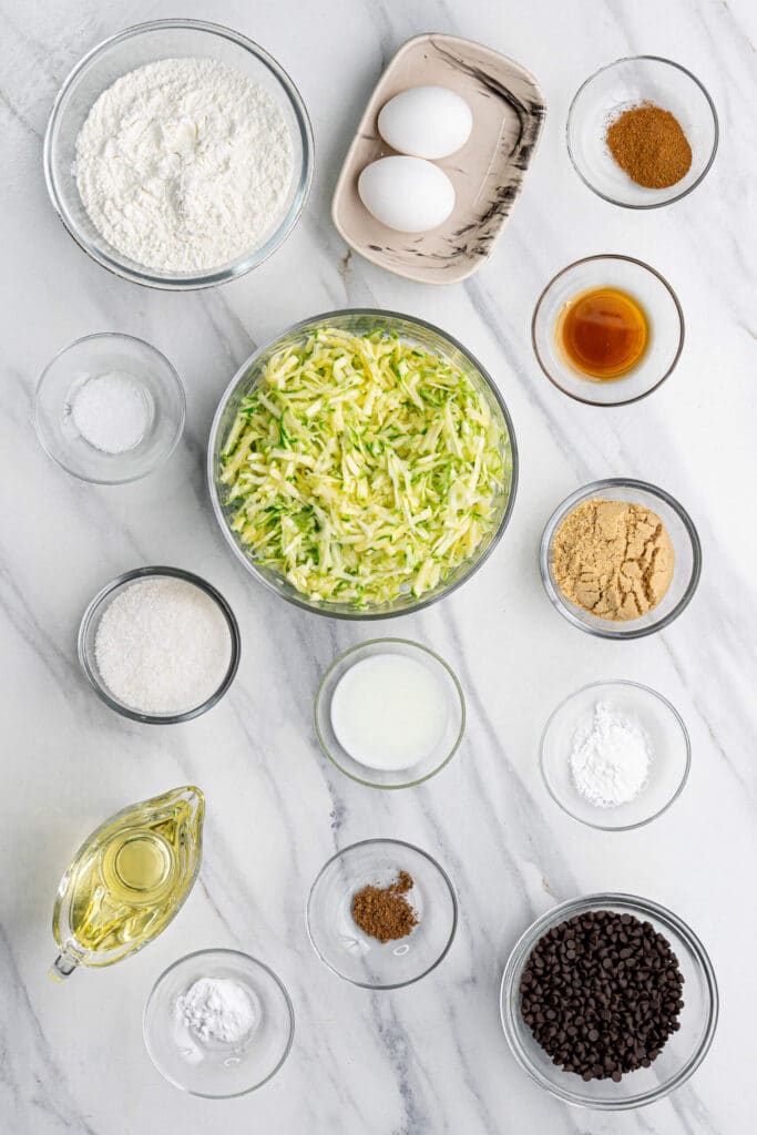 Ingredientes para hornear dispuestos sobre una superficie de mármol blanco para muffins de calabacín con chispas de chocolate, incluyendo calabacín rallado, harina, huevos, chispas de chocolate, aceite, azúcar, especias y vainilla.