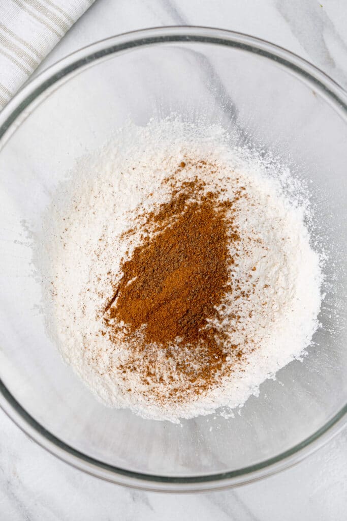 Ingredientes secos en un tazón de vidrio, incluyendo harina, canela y nuez moscada, listos para ser batidos juntos.