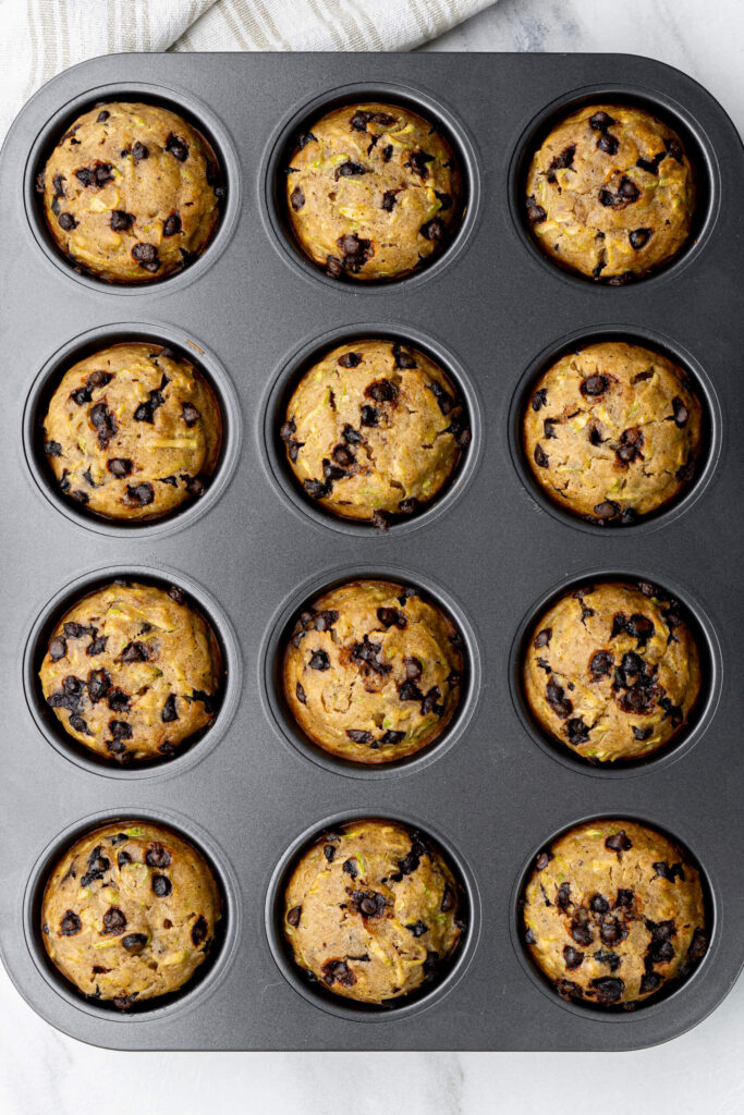 Vista aérea de muffins de calabacín con chispas de chocolate recién salidos del horno en un molde para muffins antiadherente, perfectamente levantados con bordes dorados.