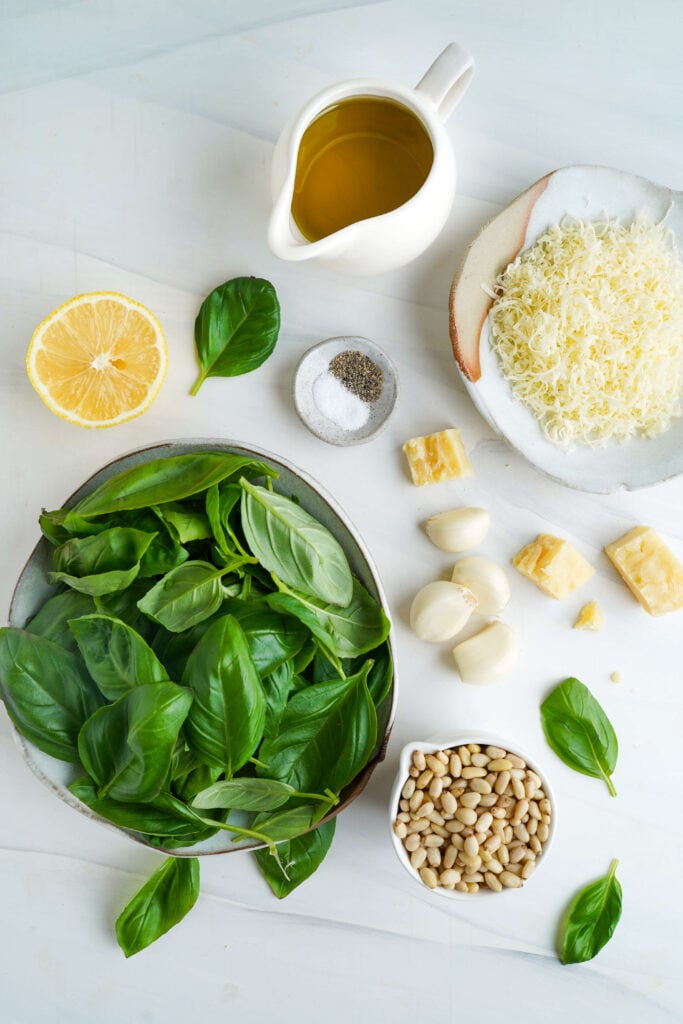 Disposición aérea de los ingredientes para el pesto de albahaca que incluye albahaca fresca, ajo, piñones, Parmesano, limón y aceite de oliva.