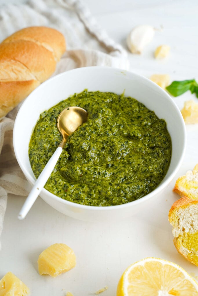 Pesto de albahaca servido en un tazón blanco con una cuchara dorada, rodeado de Parmesano, limón y pan crujiente.