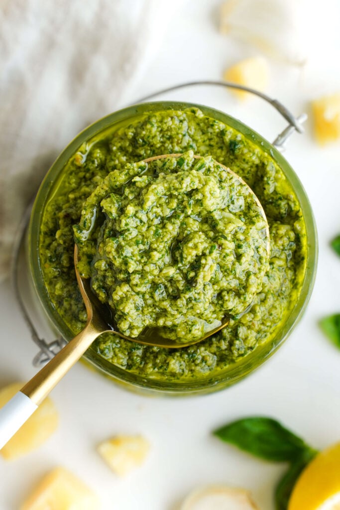 Cucharada de pesto de albahaca en un frasco de vidrio, rodeada de albahaca fresca, ajo, limón y Parmesano.