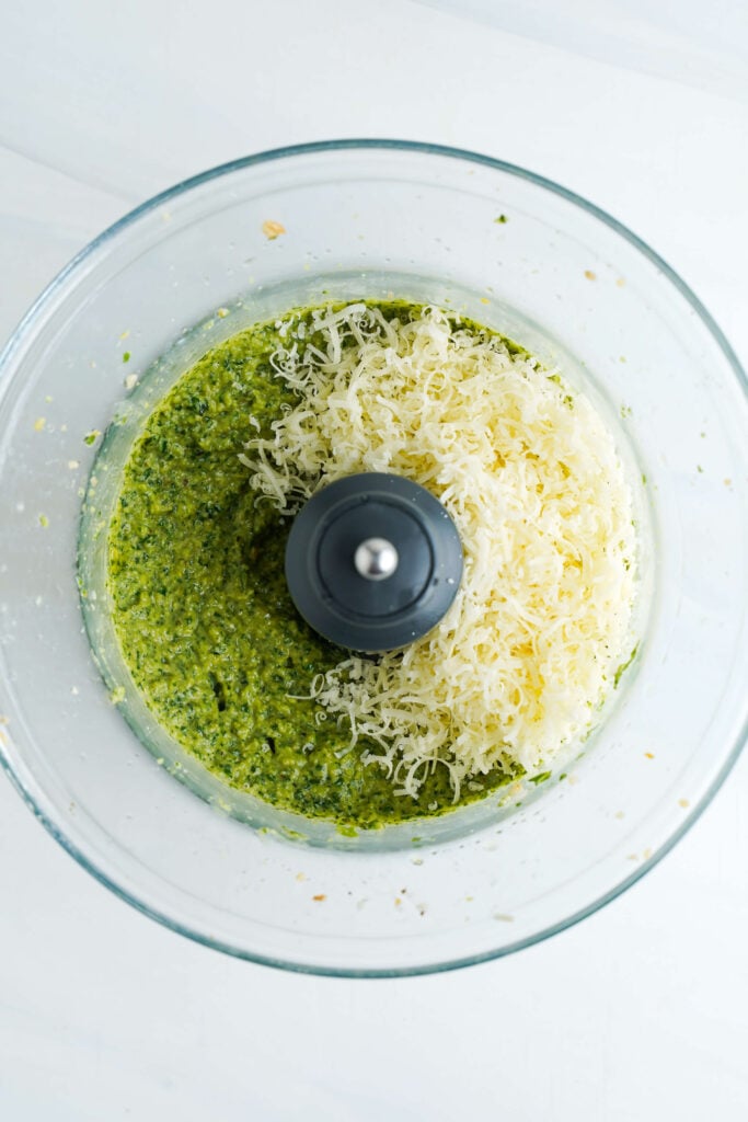 Queso Parmesano rallado recién añadido a la mezcla de pesto en un procesador de alimentos.