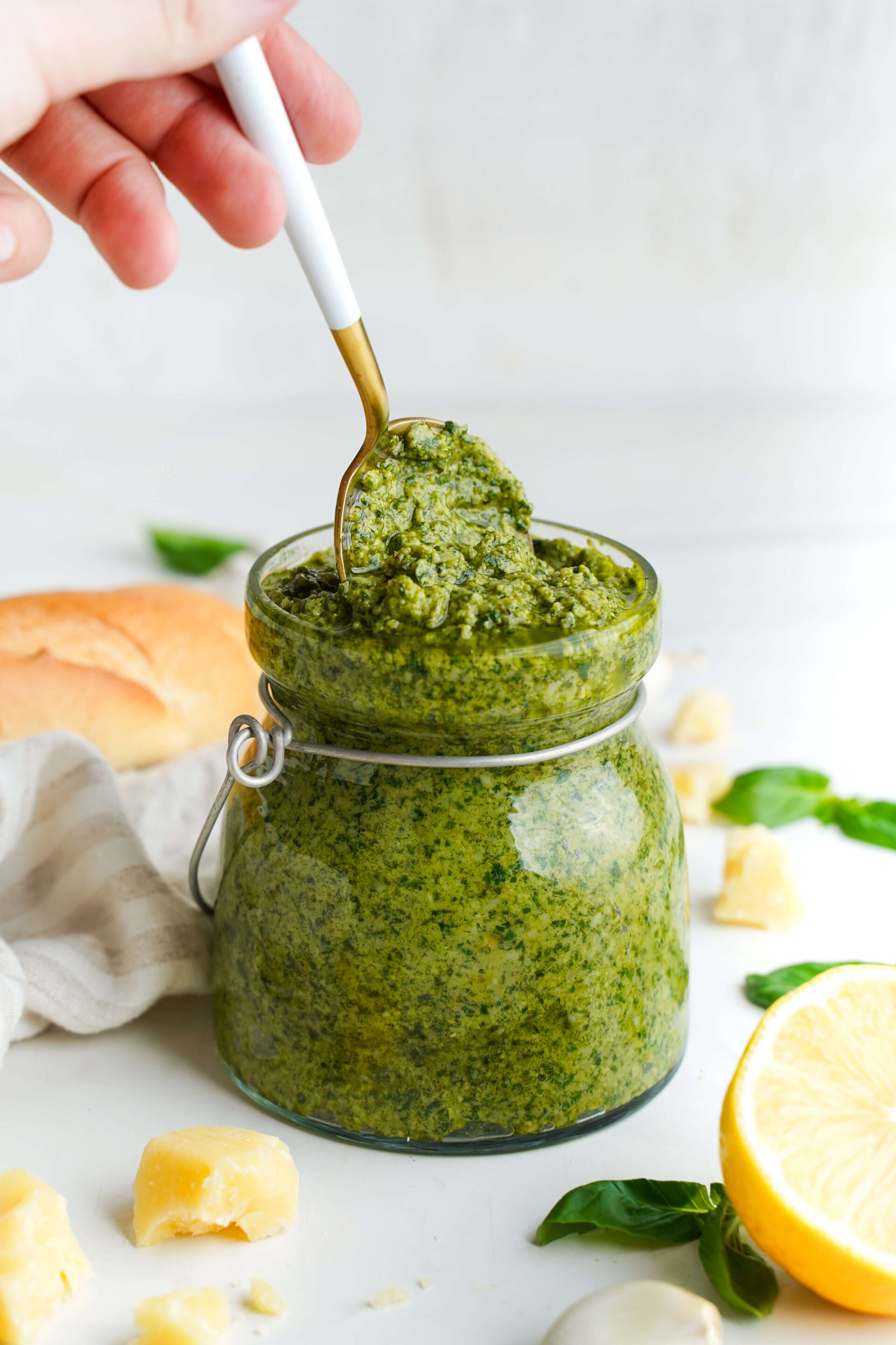 Pesto casero en un frasco de vidrio con una cuchara, rodeado de albahaca, Parmesano, limón y pan.
